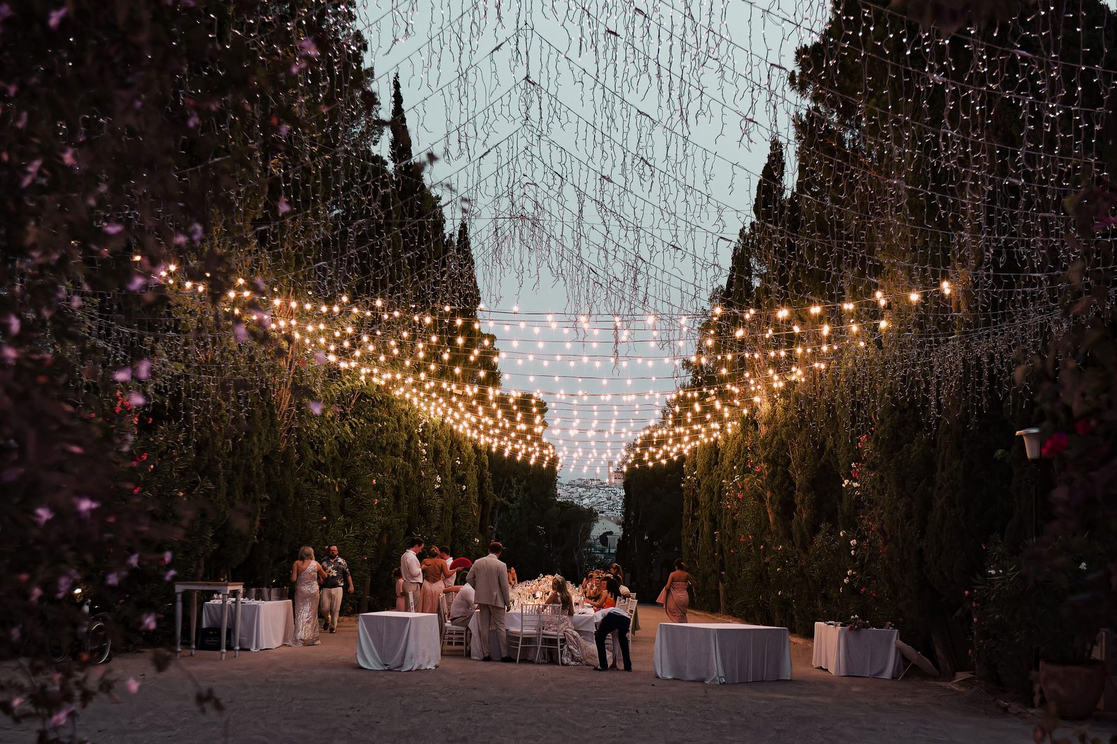 Guests gather at round tables beneath strings of lights and hanging decor between tall trees