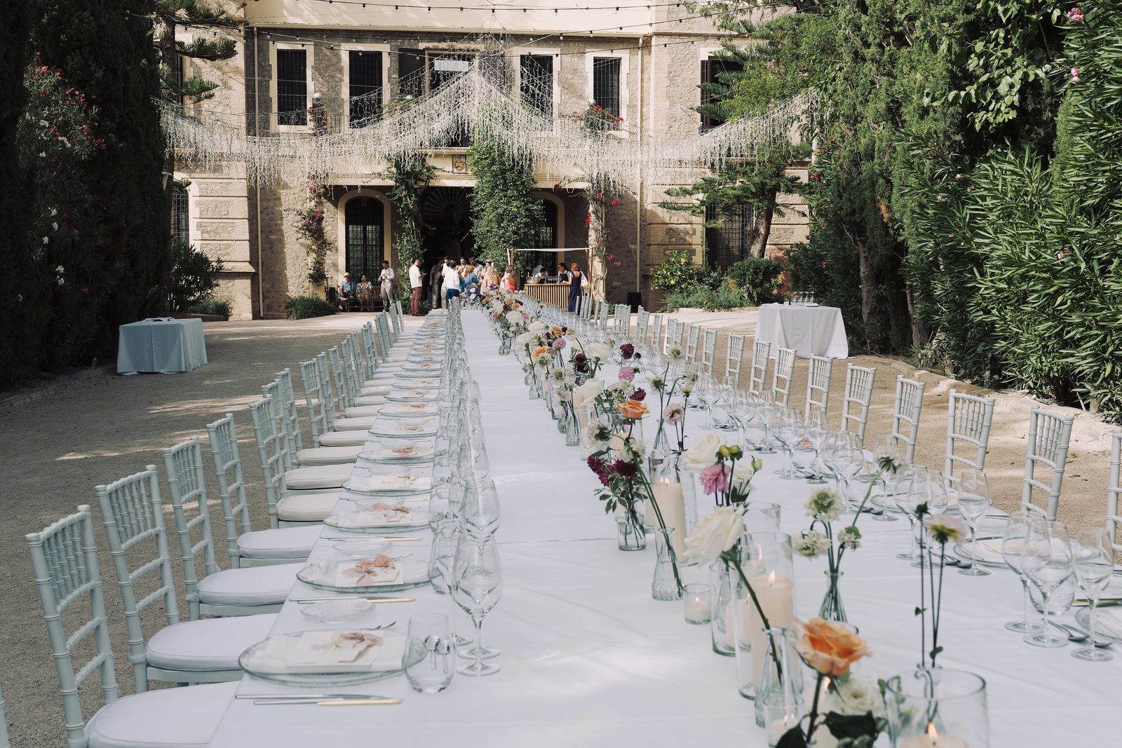 A long banquet table set with white cloth and floral centerpieces outdoors